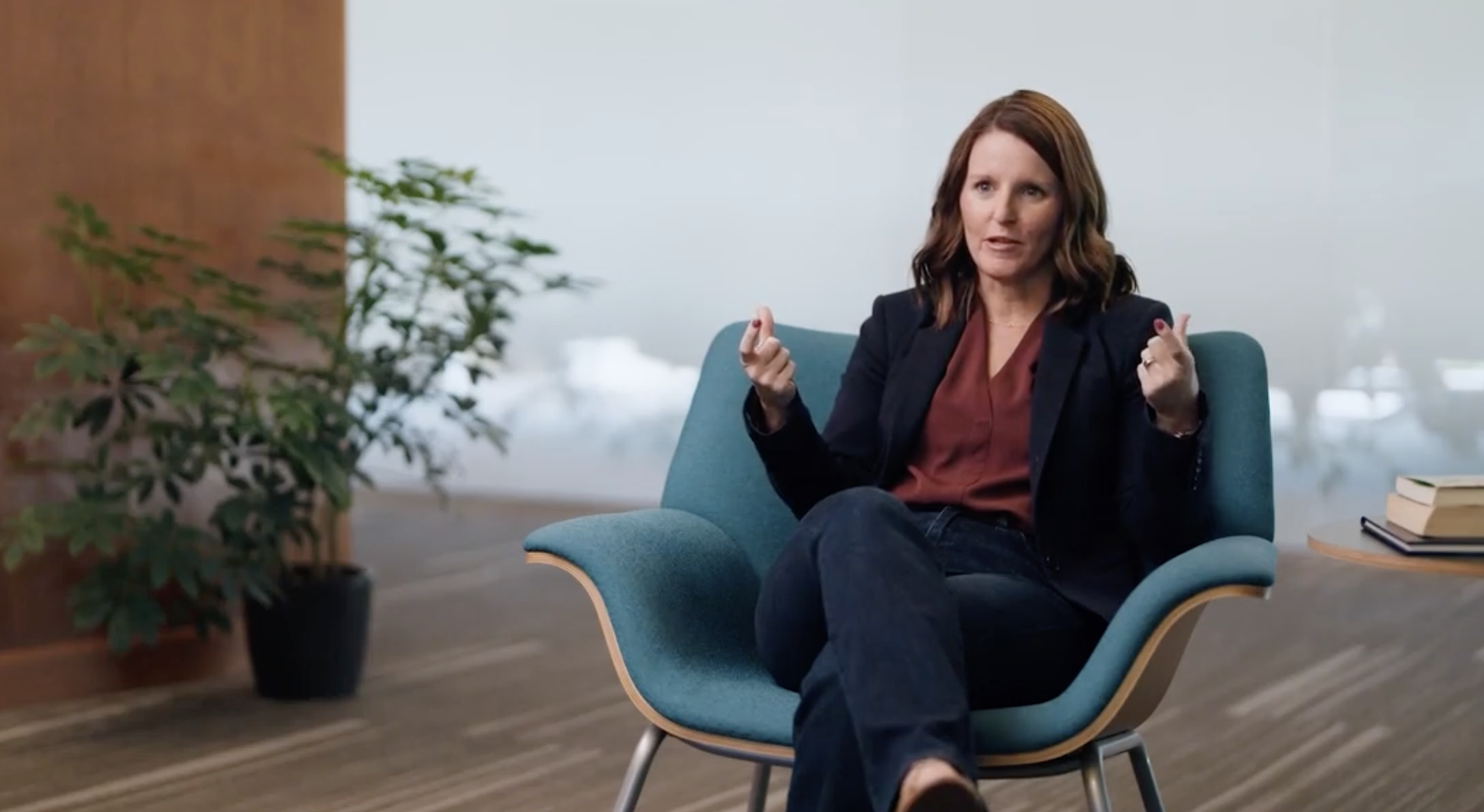 A woman sits in a modern blue chair, gesturing as she discusses ML for insurance. She wears a blazer and jeans, with a plant and a small table with books in the background.
