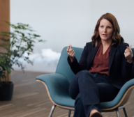 A woman in business attire sits in a modern chair, gesturing as she discusses ML for insurance. A plant and a small table with books add a professional touch to the background.