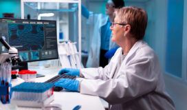 Scientist in a lab coat and gloves working at a computer in a laboratory, with scientific equipment and another researcher in the background.