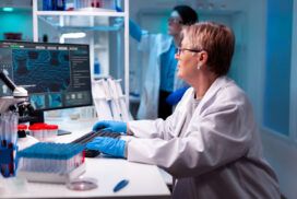 Scientist in a lab coat and gloves working at a computer in a laboratory, with scientific equipment and another researcher in the background.